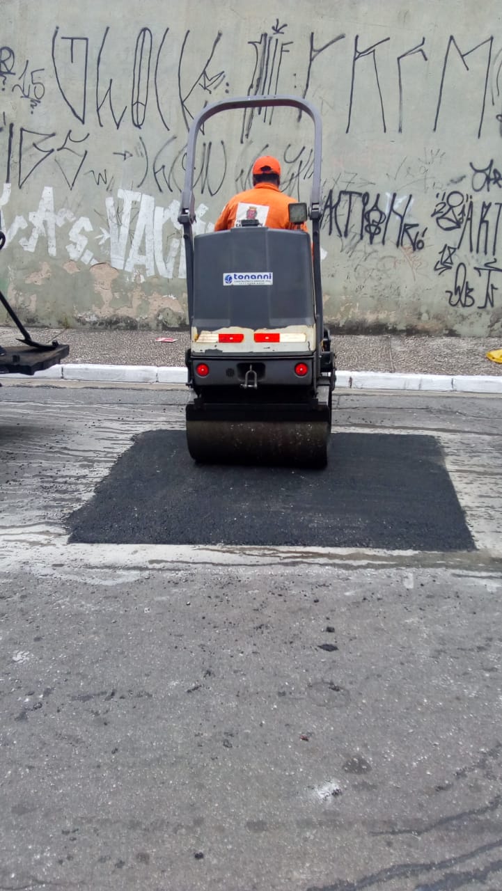 Funcionário da Prefeitura trajado de uniforme laranja finaliza o asfaltamento de um buraco, utilizando um rolo compressor motorizado.